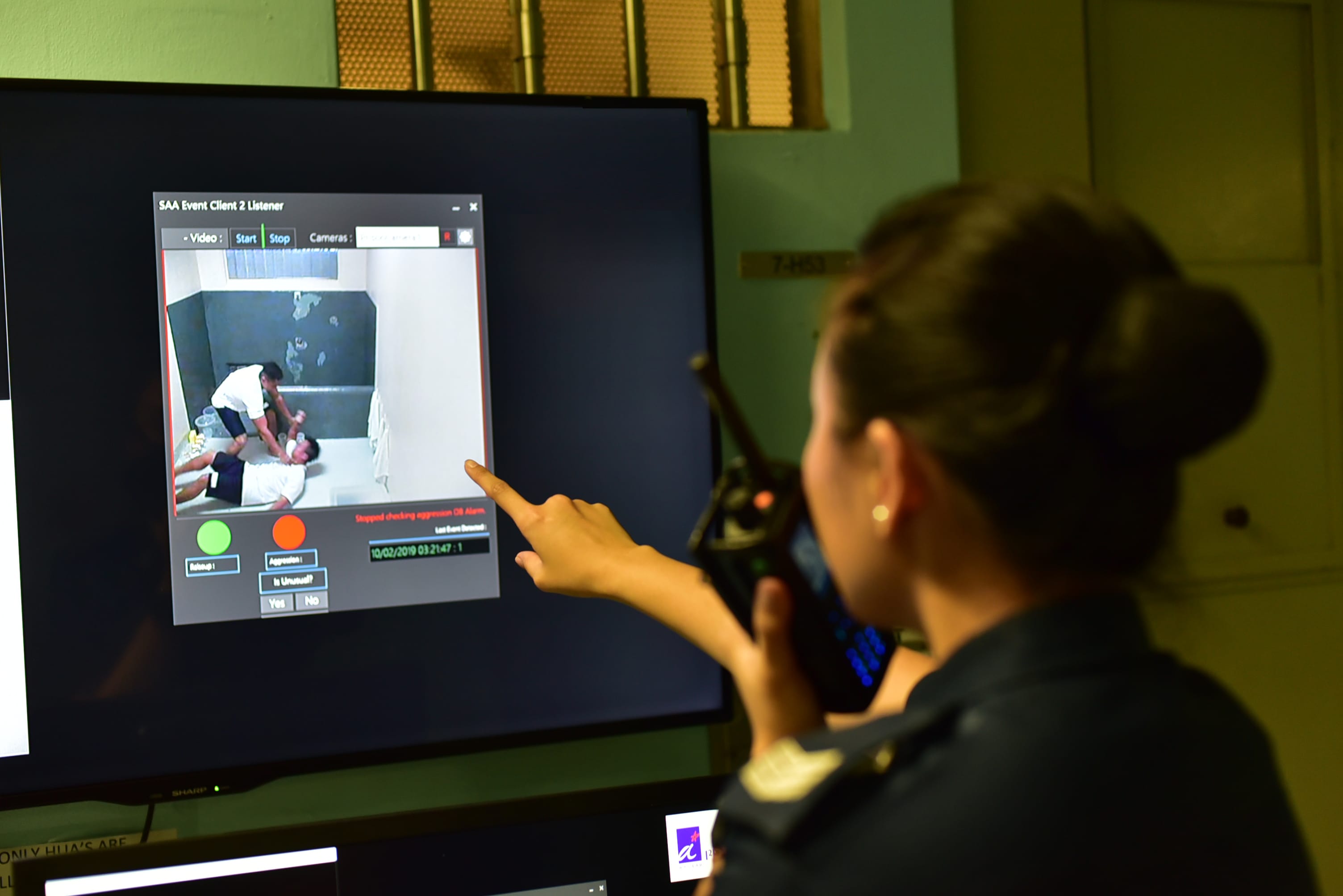 A woman points at a monitor displaying a security footage of an altercation between two people in a small room.